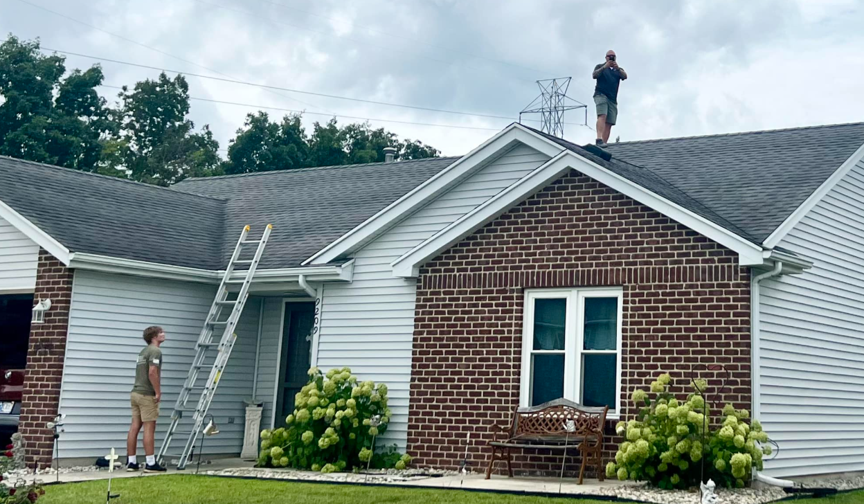Two workers on a house roof with ladders and a brick-front home below.