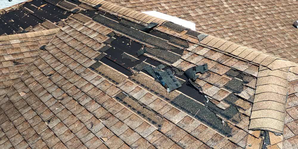A high-angle view shows a damaged section of a brown shingled roof with missing shingles, exposing the black underlayment.