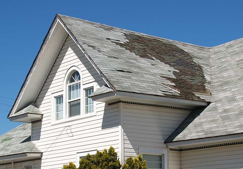 House roof with extensive weathered and peeling shingles on a white exterior under a blue sky