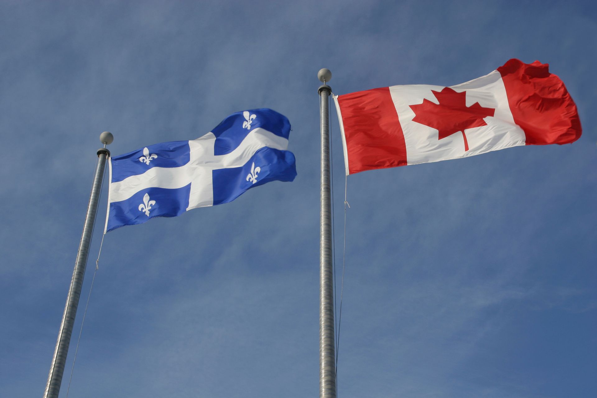 Les drapeaux du Québec et du Canada flottent sur des mâts se détachant sur un ciel bleu.
