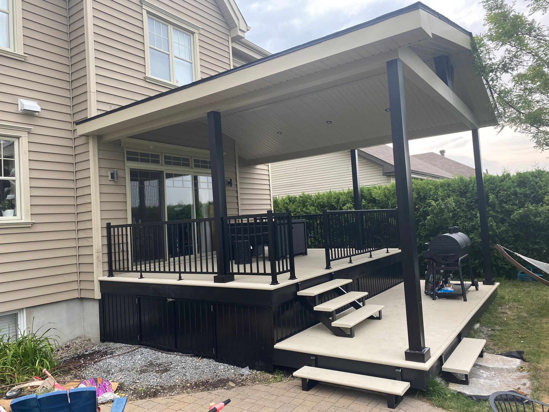Terrasse couverte à l'arrière d'une maison beige, avec escalier, rambarde et mobilier de jardin.