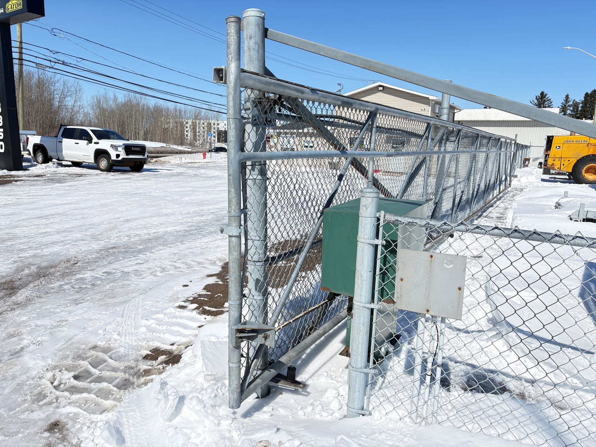 Portail en grillage dans une zone industrielle enneigée, avec des camions stationnés et une chargeuse jaune en arrière-plan.
