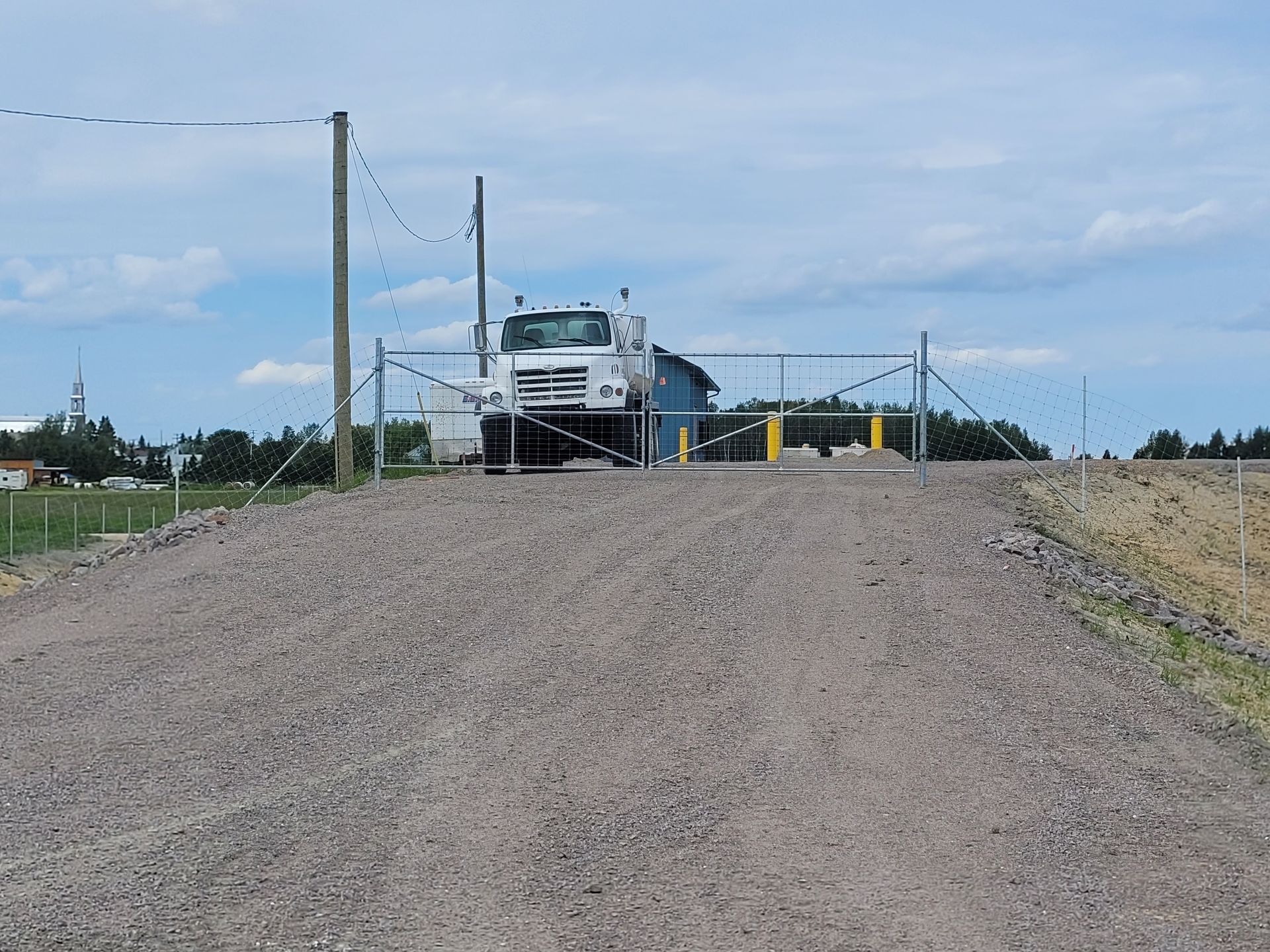 Chemin de gravier menant à un site industriel clôturé, avec des camions et du matériel sous un ciel nuageux.