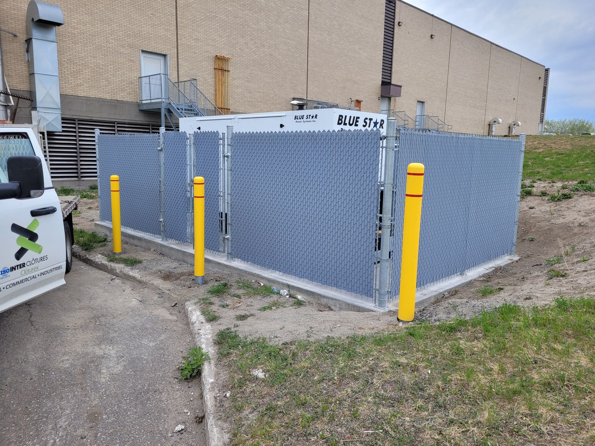 Bornes jaunes autour d'un boîtier technique gris à côté d'un bâtiment sur un terrain gravillonné