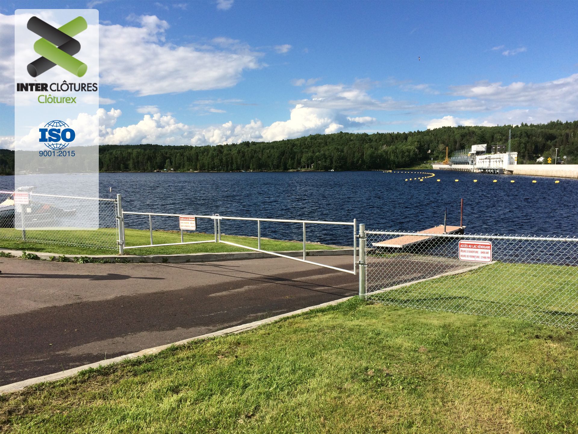 Vue sur le lac avec quai, rive herbeuse, clôture en grillage et ciel bleu nuageux