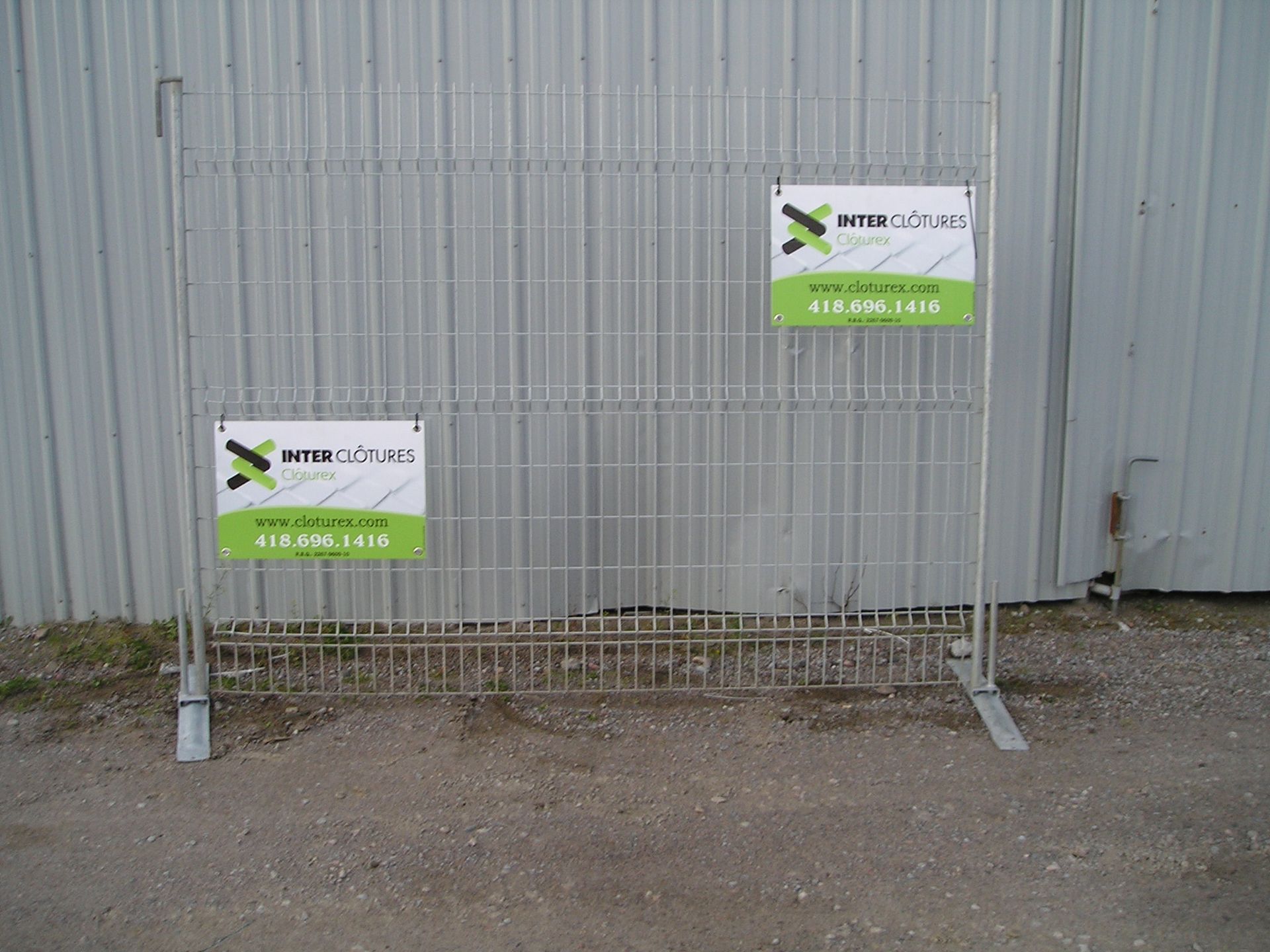 Barrière métallique avec deux panneaux contre un mur ondulé gris sur une surface de gravier.