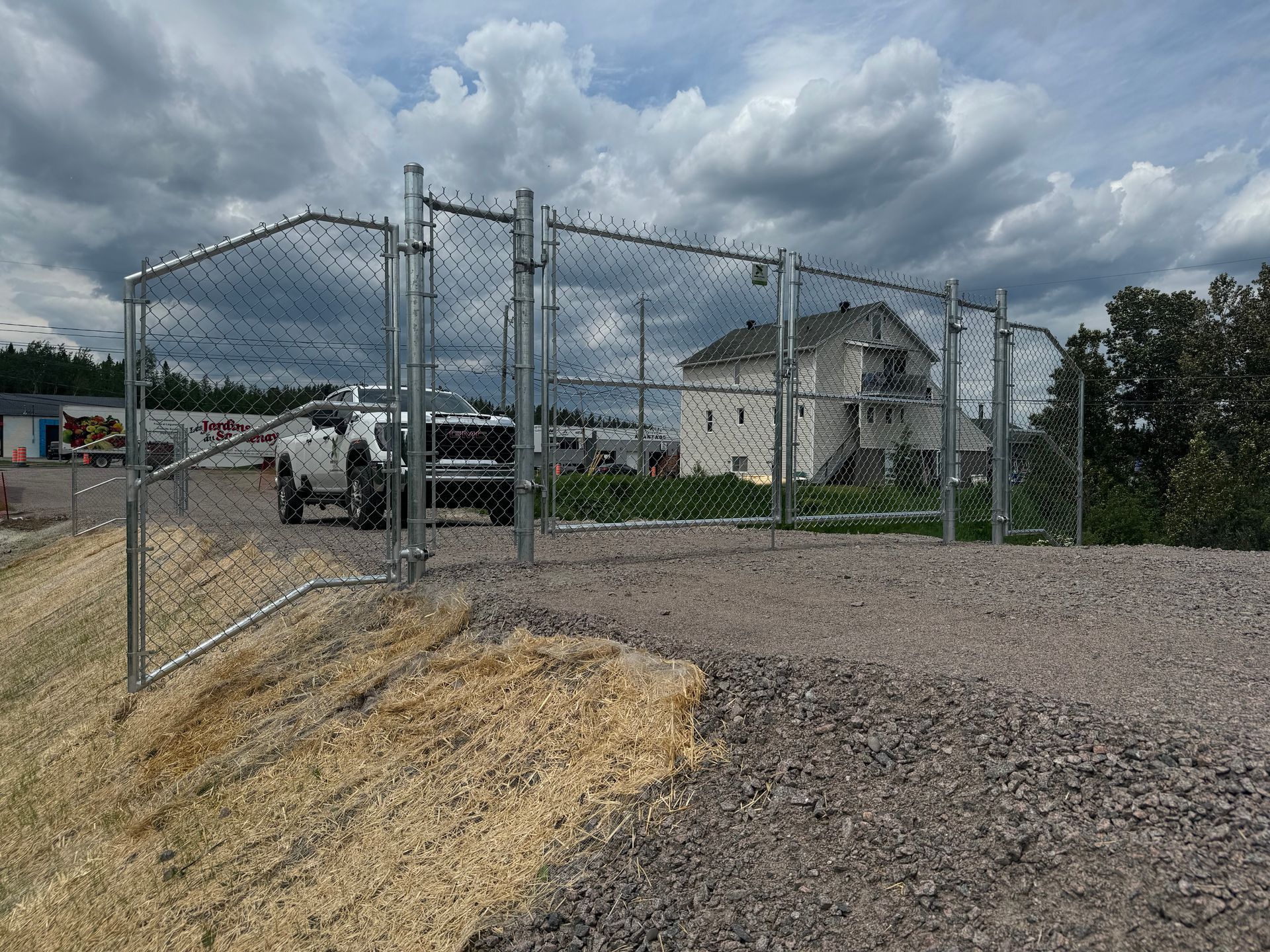 Clôture en grillage entourant un terrain gravillonné, avec une maison blanche et un ciel nuageux en arrière-plan.