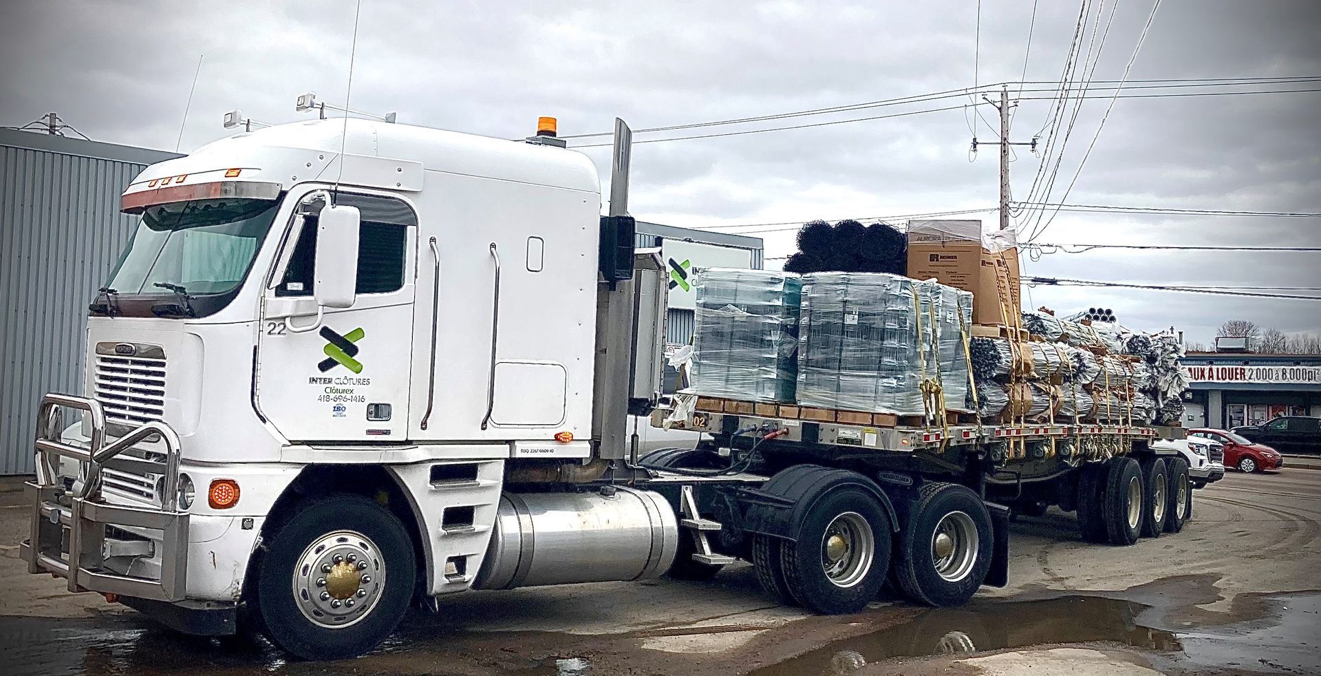 Un semi-remorque blanc transporte des marchandises empilées sur une remorque dans une zone industrielle enneigée.