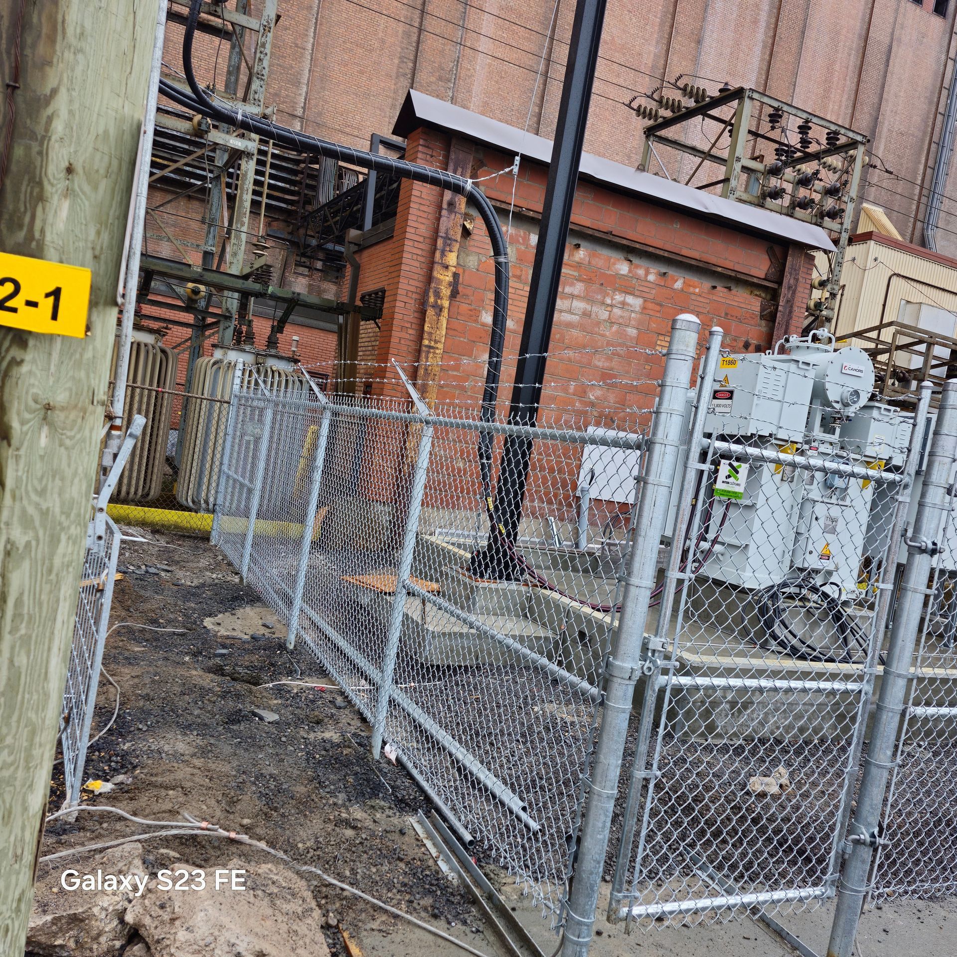 Clôture en grillage autour des équipements électriques jouxtant un bâtiment en briques et une ruelle étroite