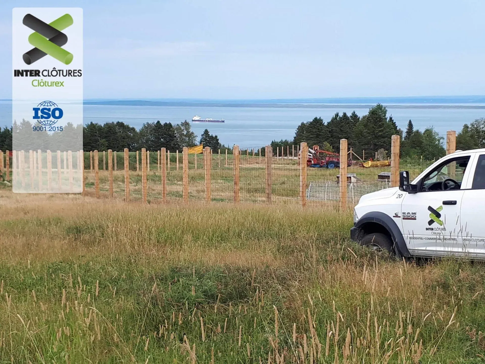 SUV blanc près d'un champ clôturé avec des poteaux électriques, de l'herbe au premier plan et un lac ou la mer en arrière-plan.