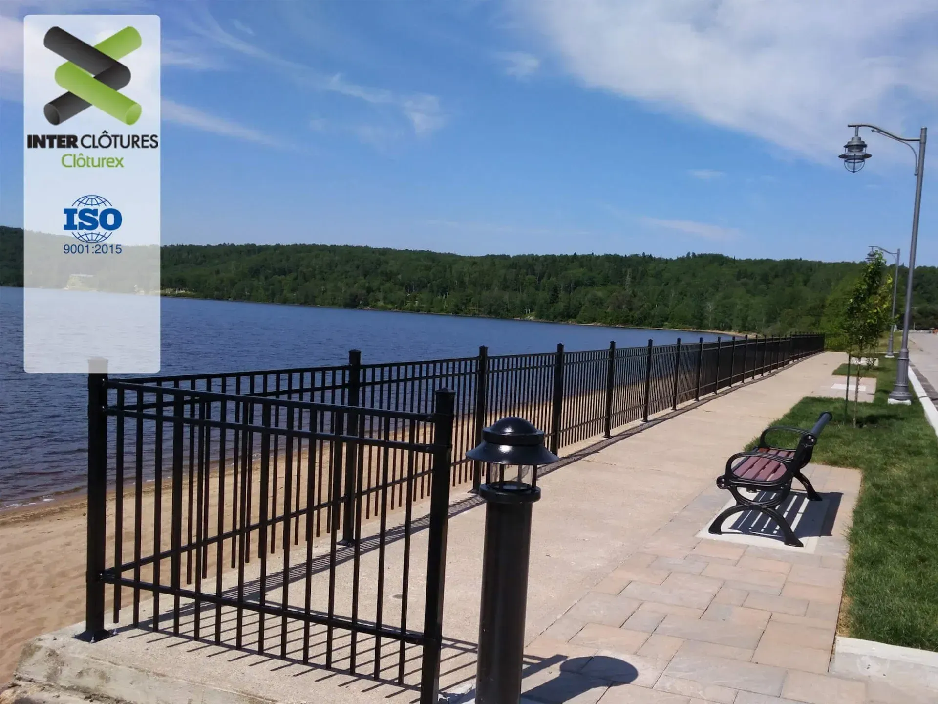 Promenade au bord de la rivière avec rambardes noires, bancs et lampadaire au bord d'une eau calme sous un ciel bleu