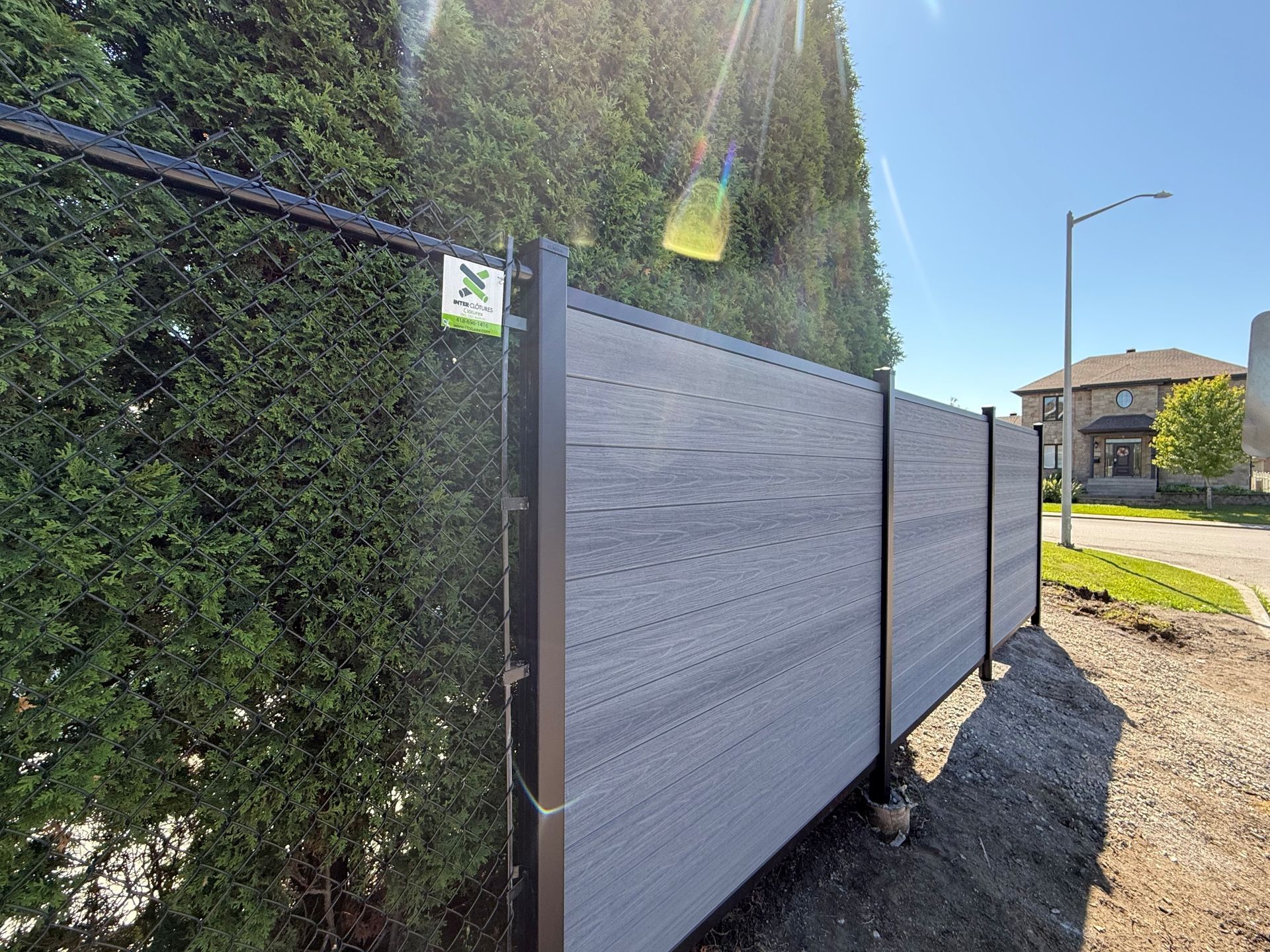 Des panneaux de clôture en métal gris bordent une haie le long d'une route ensoleillée.