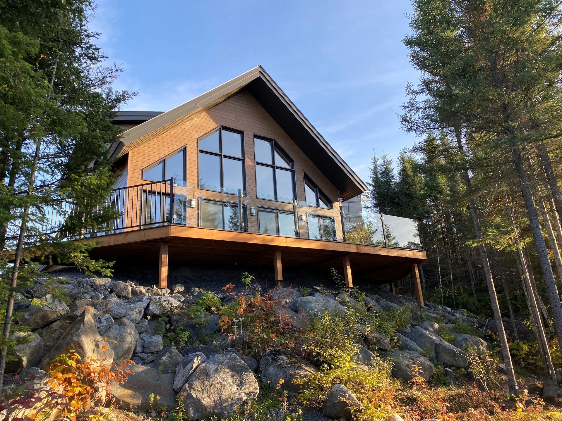 Chalet moderne sur un flanc de colline rocailleux, entouré de pins, avec de grandes fenêtres et une terrasse en bois.