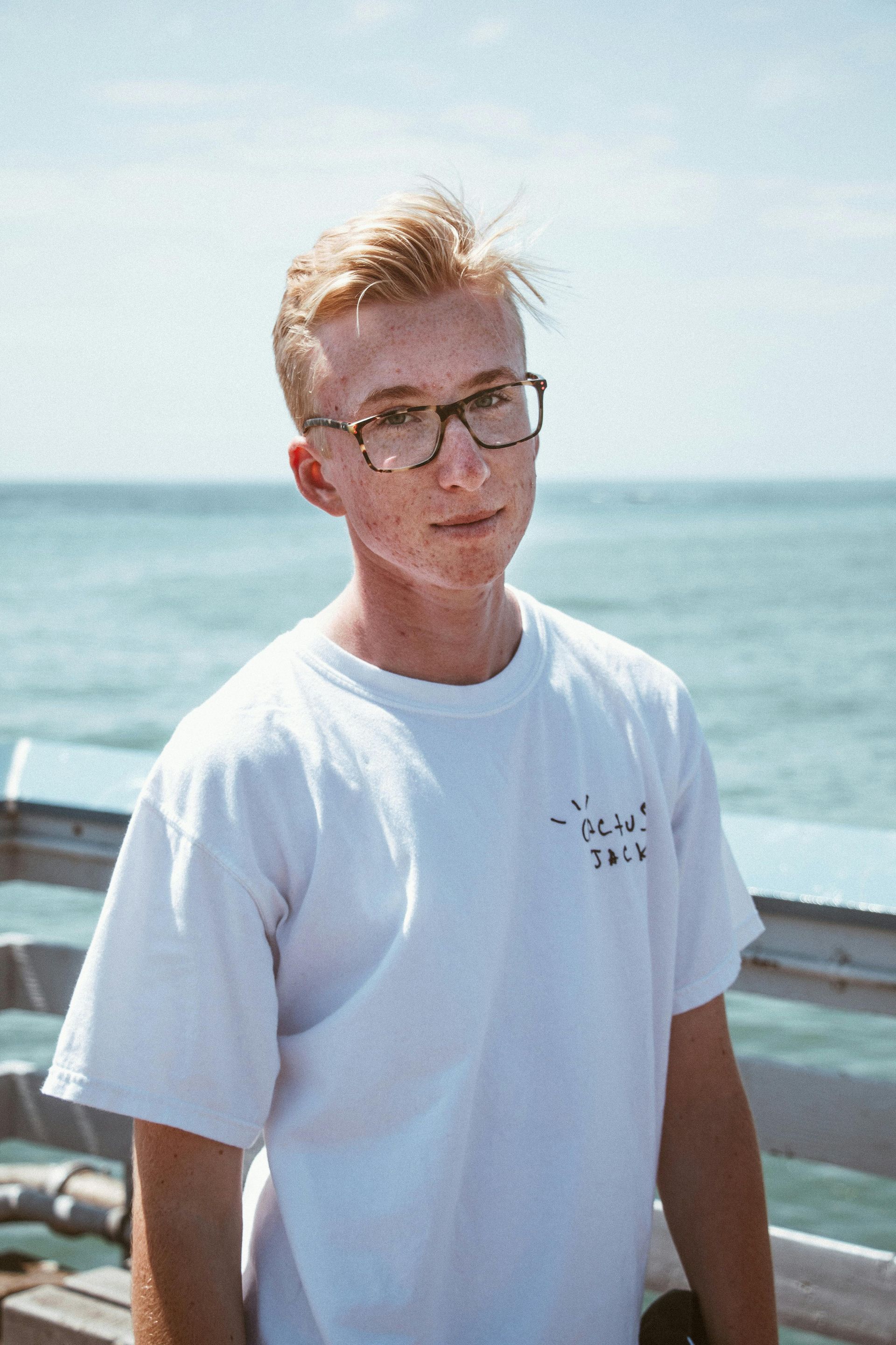 Person in a white T-shirt standing by the ocean, with a boat railing behind them.
