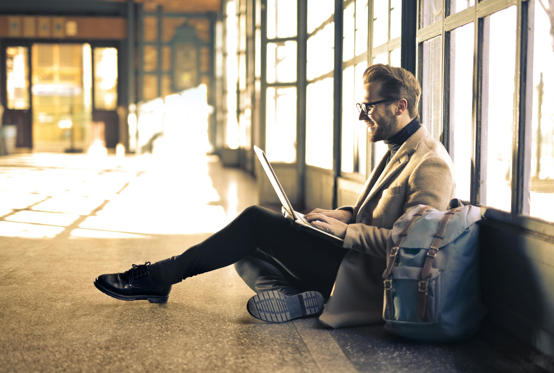 Man sitting on the floor by windows, using a laptop beside a gray backpack in a bright hallway