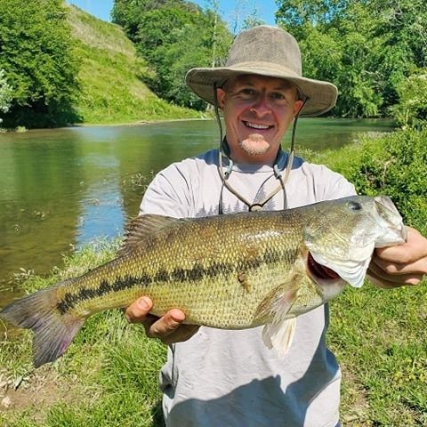 a man in a hat is holding a large fish in front of a river .