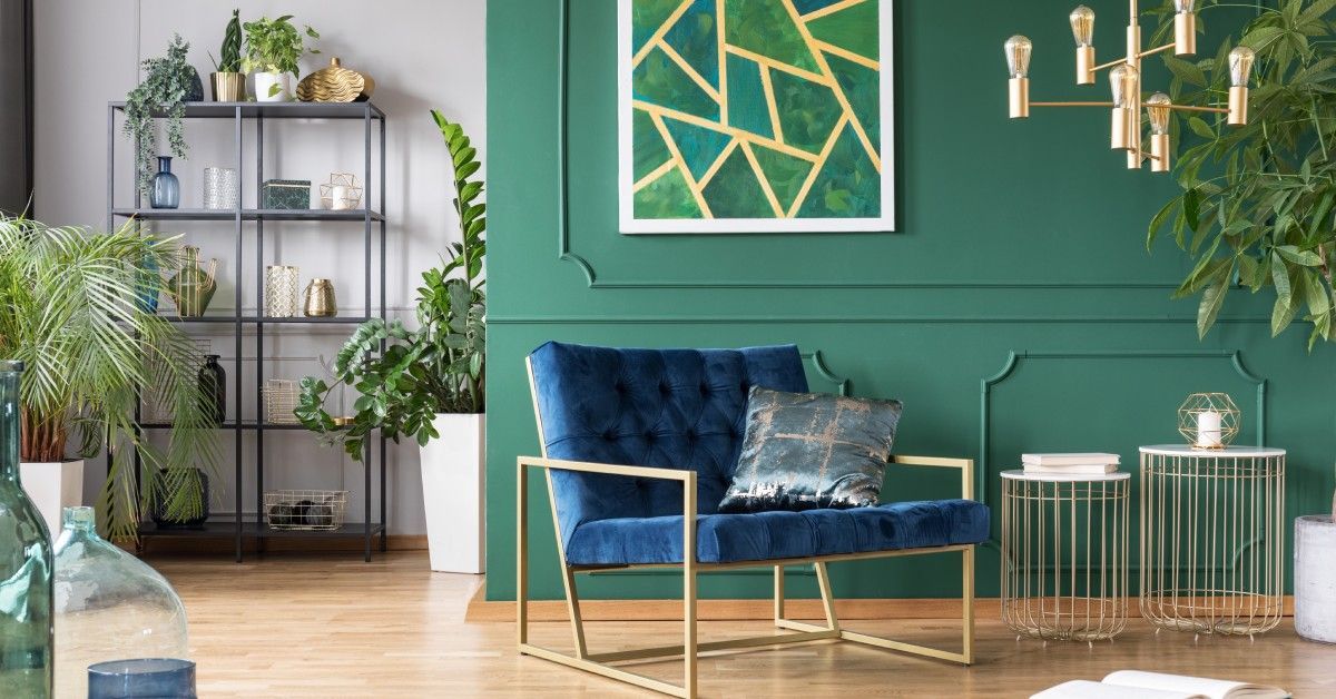 A room has a green wall with trim and a framed abstract painting. A wire shelf with plants and vases is in the background.