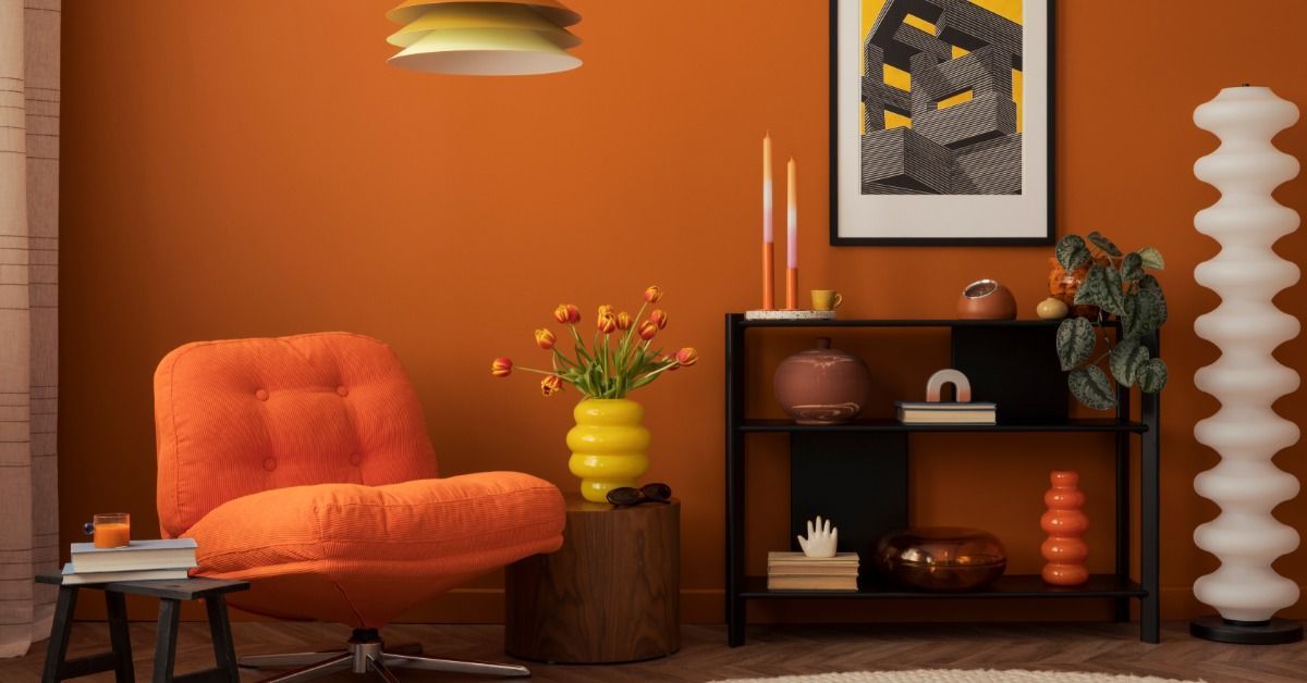 A nook has an orange chair, dark orange walls, a black shelf with plants and vases, and a framed piece of art on the wall.
