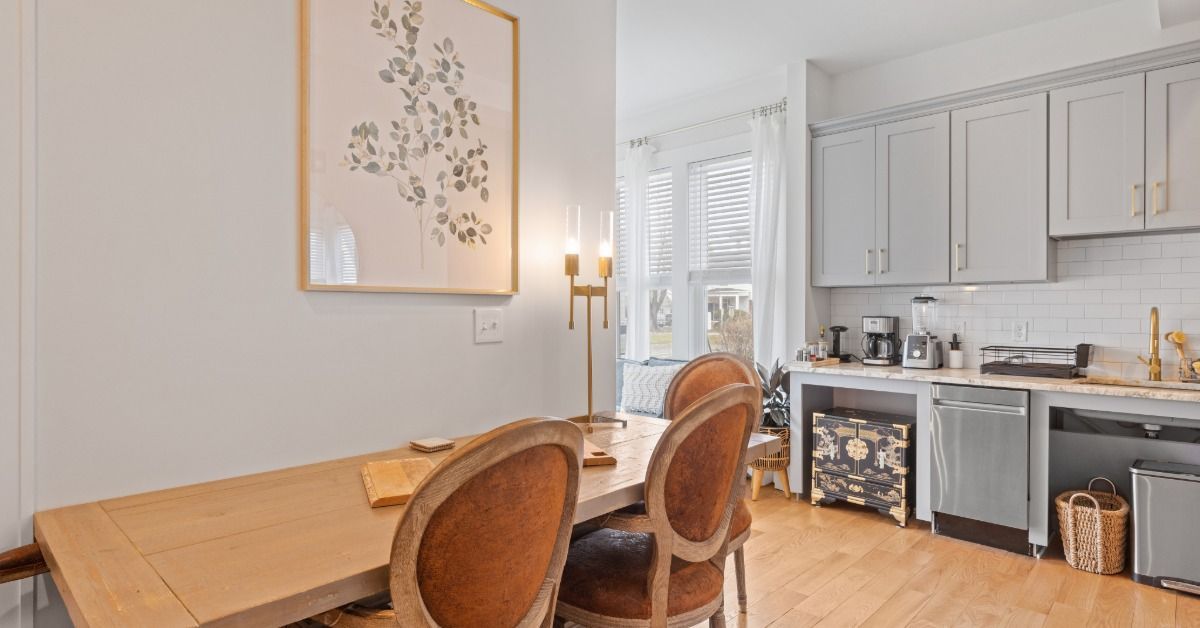 A light wood table with chairs are against a wall to the left of the kitchen. A framed floral art print is on the wall.