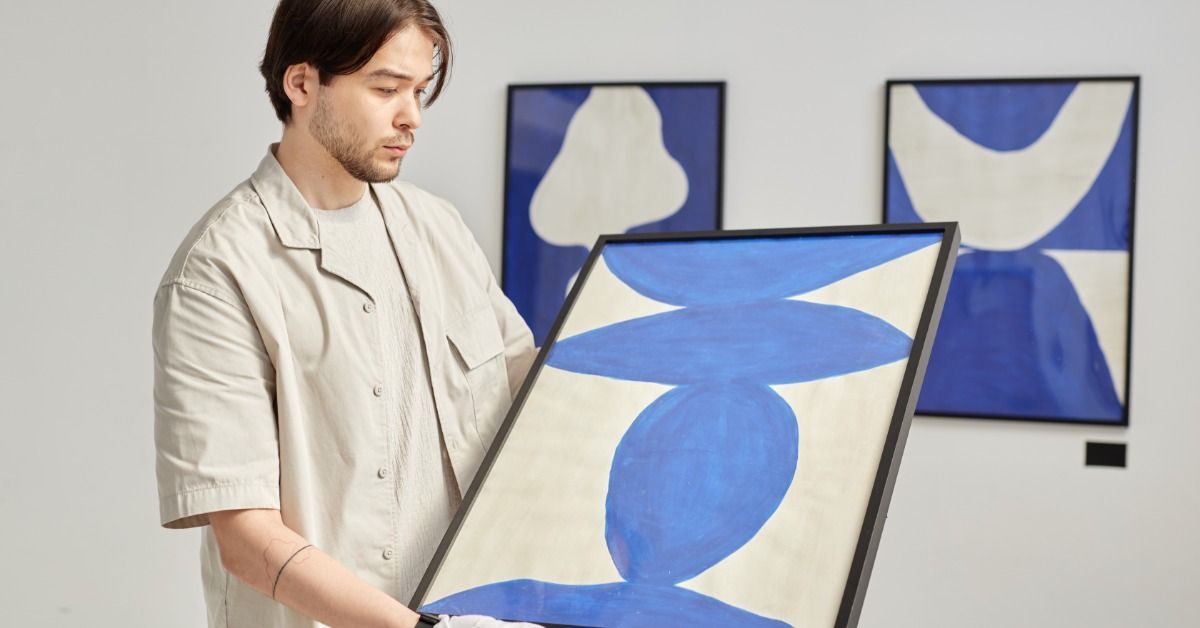 A man wearing white gloves holds a piece of abstract art in a black frame.
