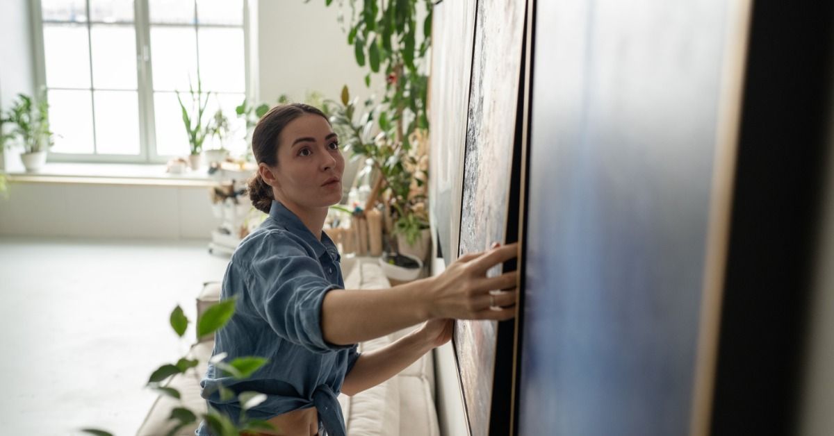 A woman carefully adjusts a framed piece of art onto a wall. The room has natural light and several potted plants.