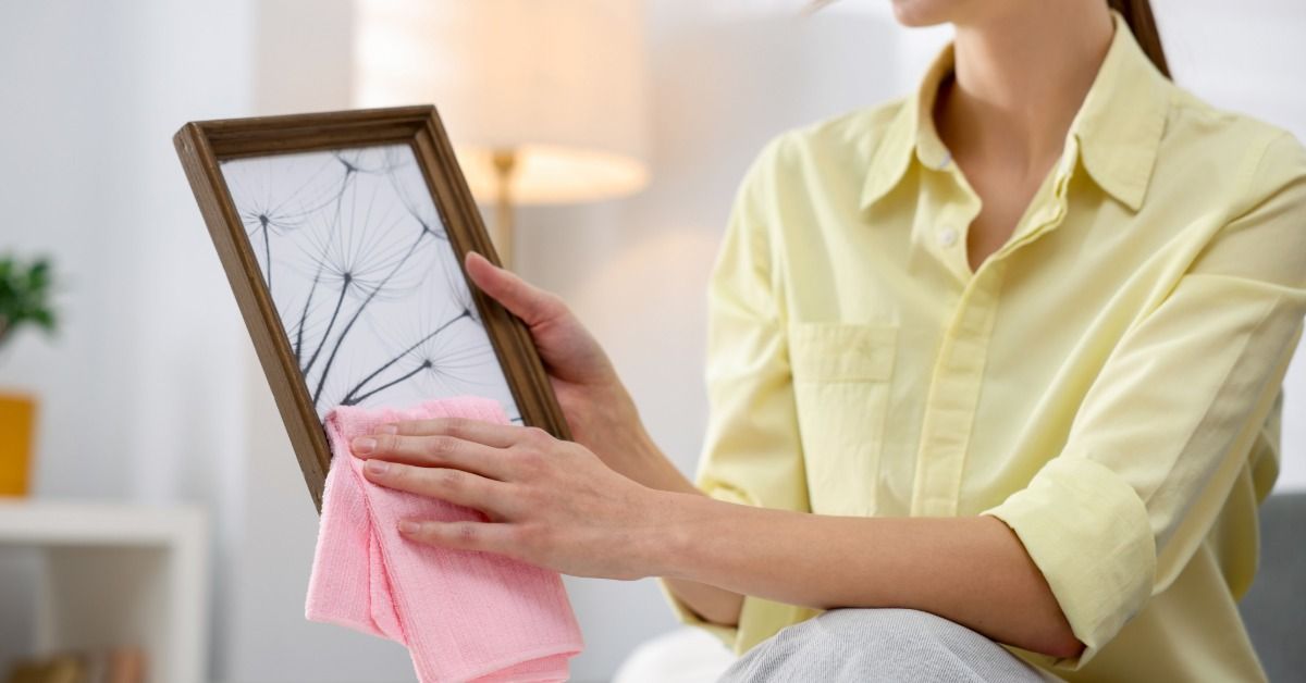 A woman is sitting as she uses a pink cloth to wipe the edges of a framed art piece. A lamp glows in the background.