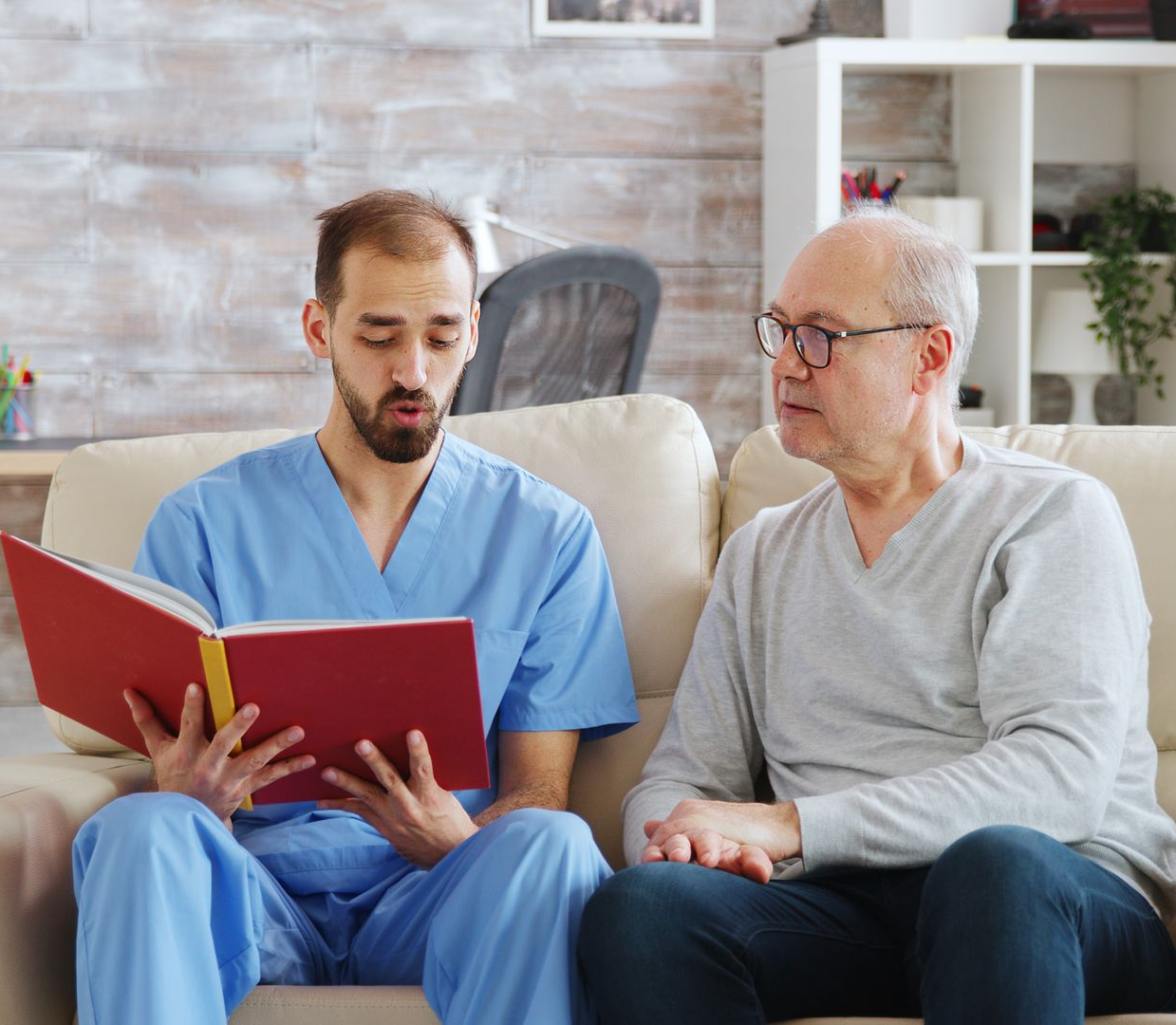 Caregiver in blue scrubs reads to older person on a couch. Crutch beside them.