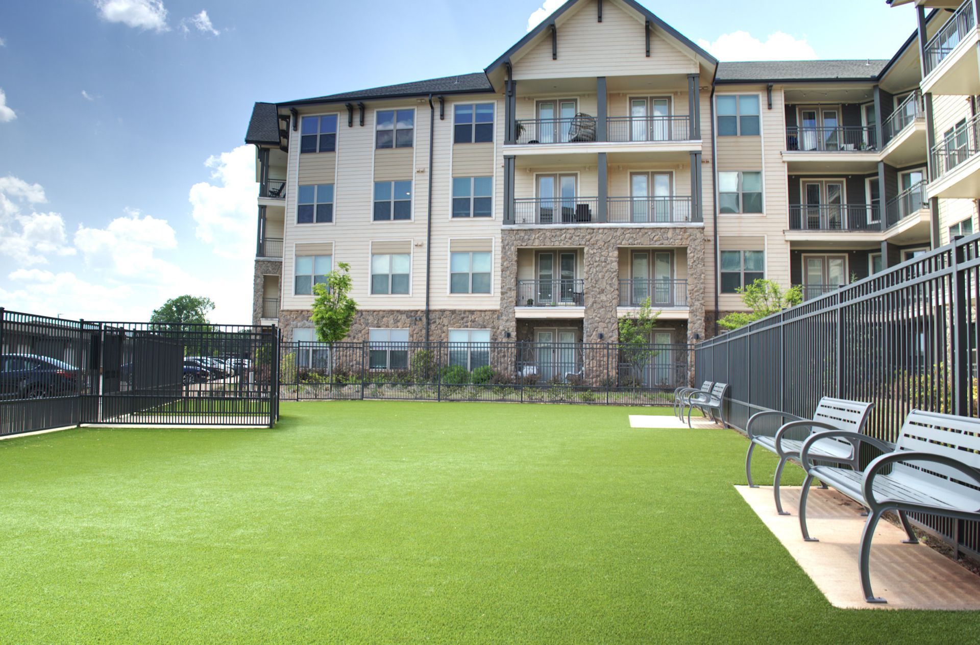 Apartment courtyard with green lawn, black fence, and benches beside a multi-story building
