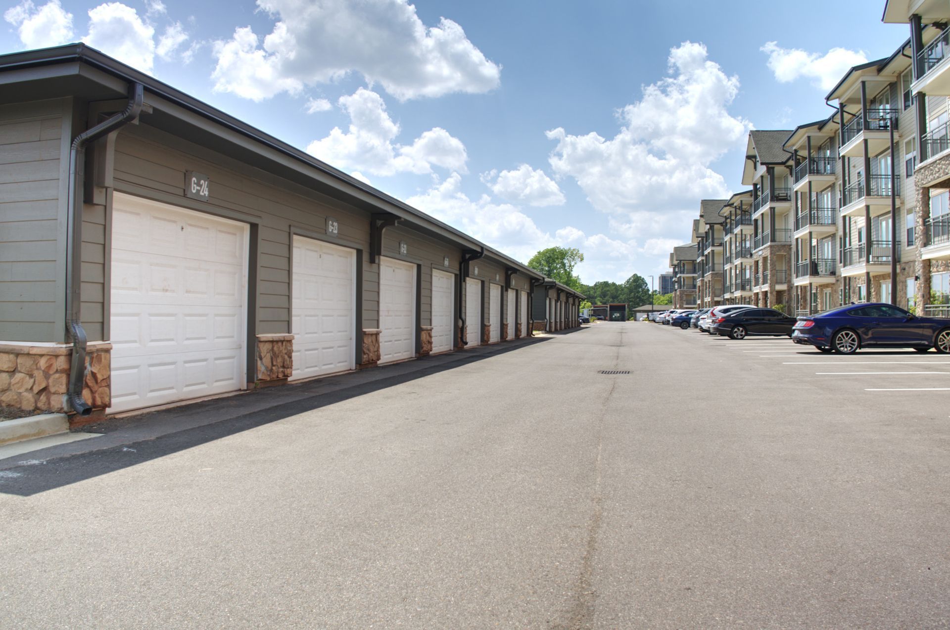 Apartment complex alley with garage doors on the left and parked cars along the right under a partly cloudy sky