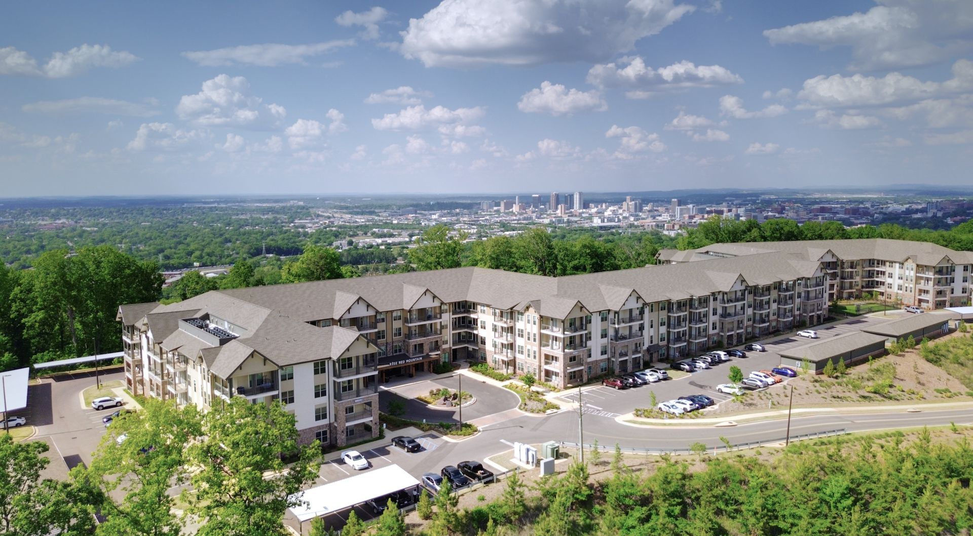 Apartment complex with gray roofs and parking lot, overlooking a distant city skyline on a sunny day