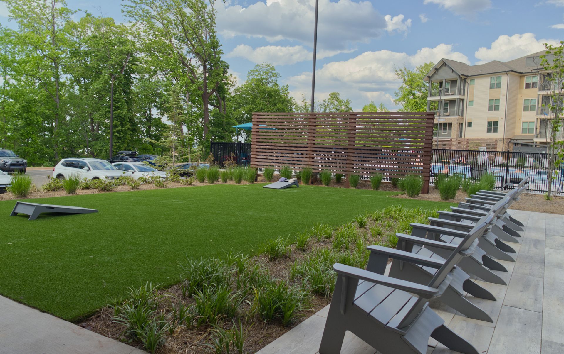 Outdoor courtyard with lounge chairs, green lawn, trees, and apartment buildings under a partly cloudy sky