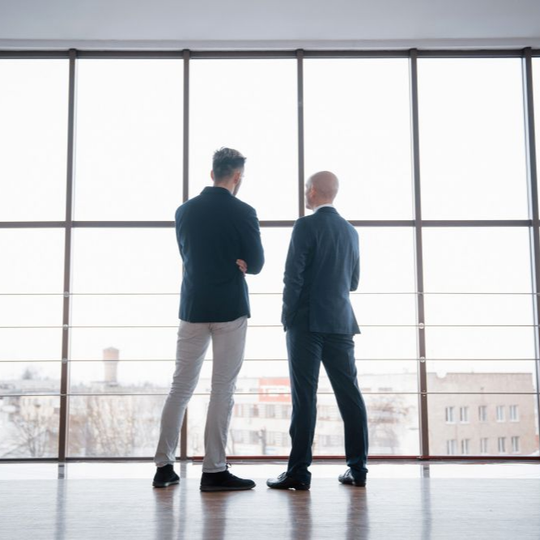 Two people in suits standing by large windows, looking out over a city skyline.