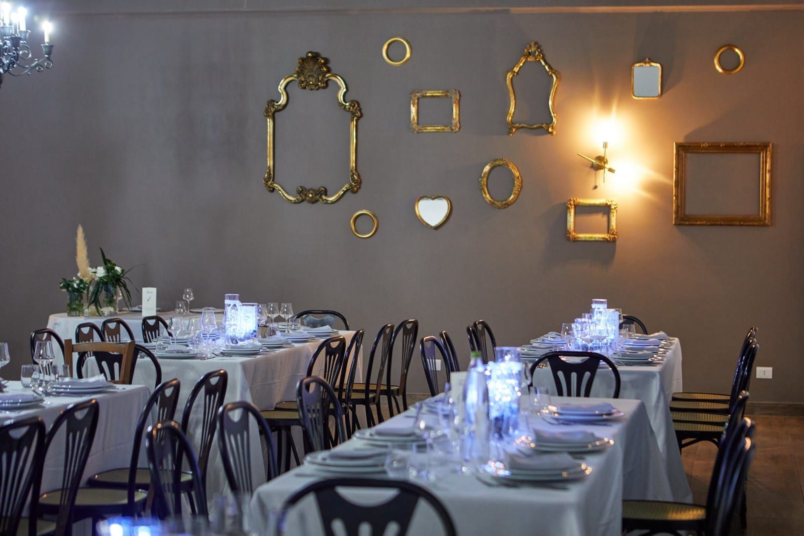 Sala da pranzo del ristorante con tavoli apparecchiati per una festa. Cornici dorate adornano una parete grigia.
