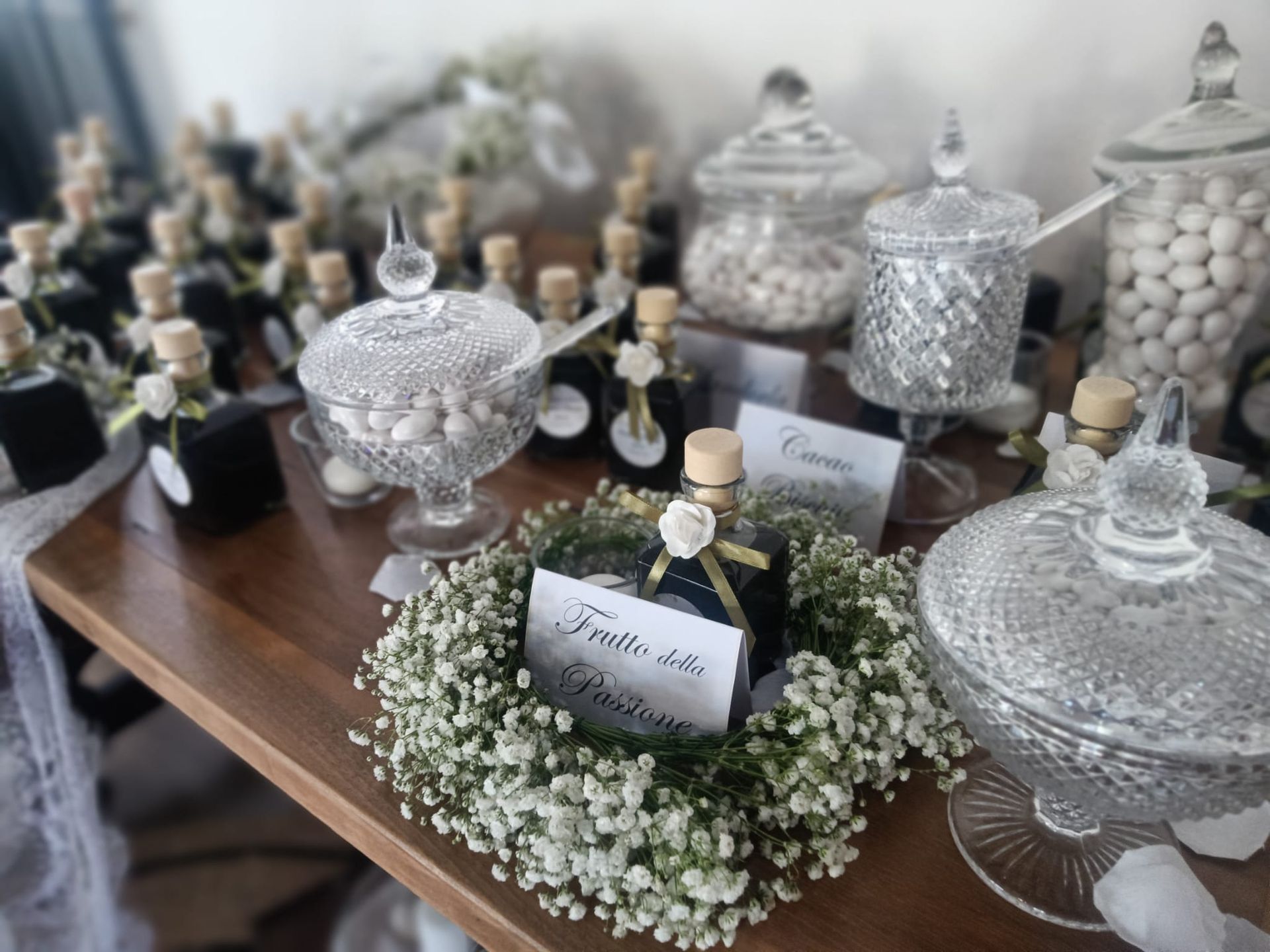 Tavolo delle bomboniere: barattoli di caramelle, bottiglie di olio d'oliva e ghirlande di fiori su una superficie di legno.