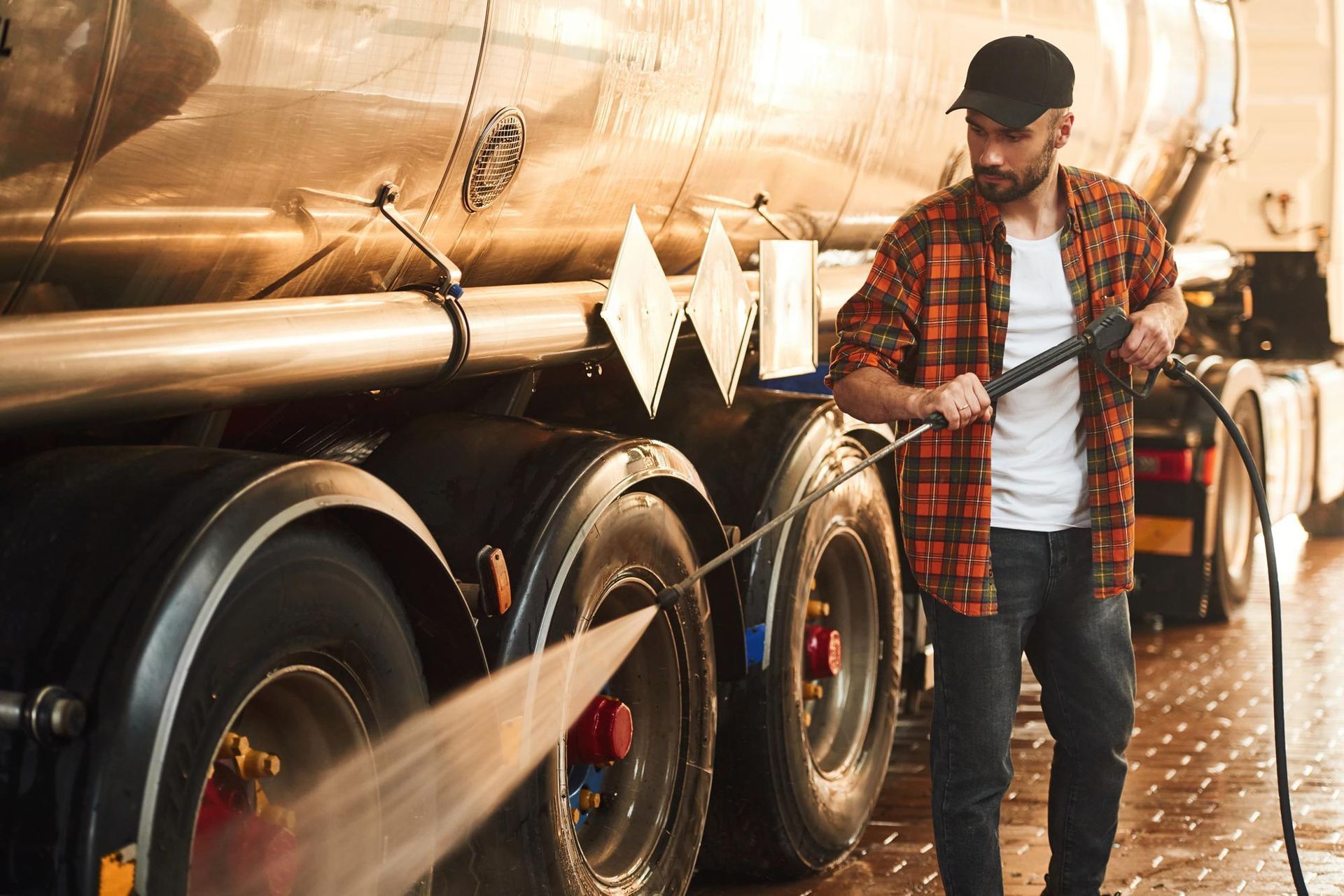 Man power washing a truck's tires with a hose, wearing a plaid shirt and a black cap, outdoors.