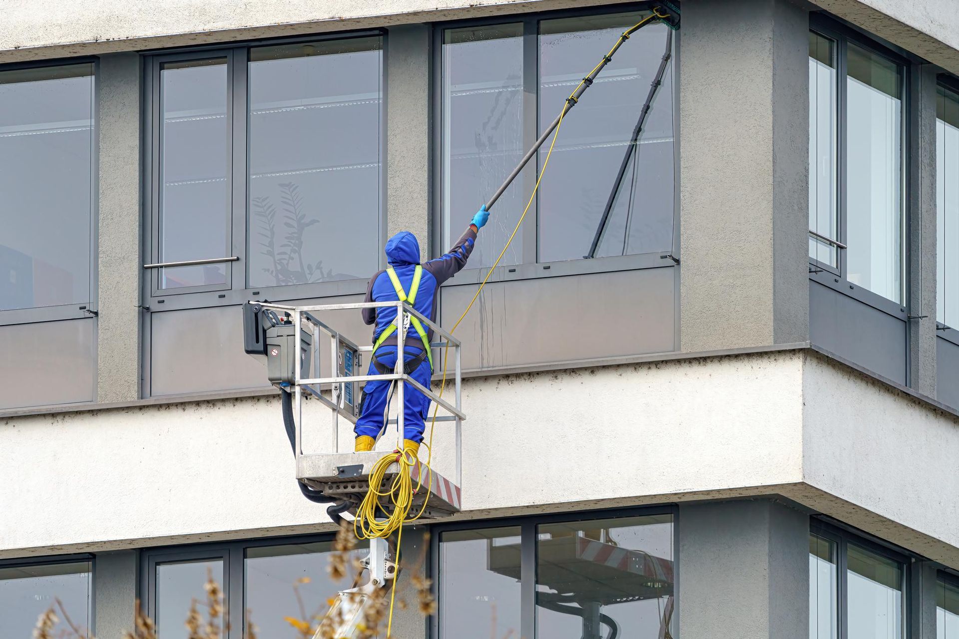 Window washer in blue overalls on a lift, cleaning building windows with an extended pole.