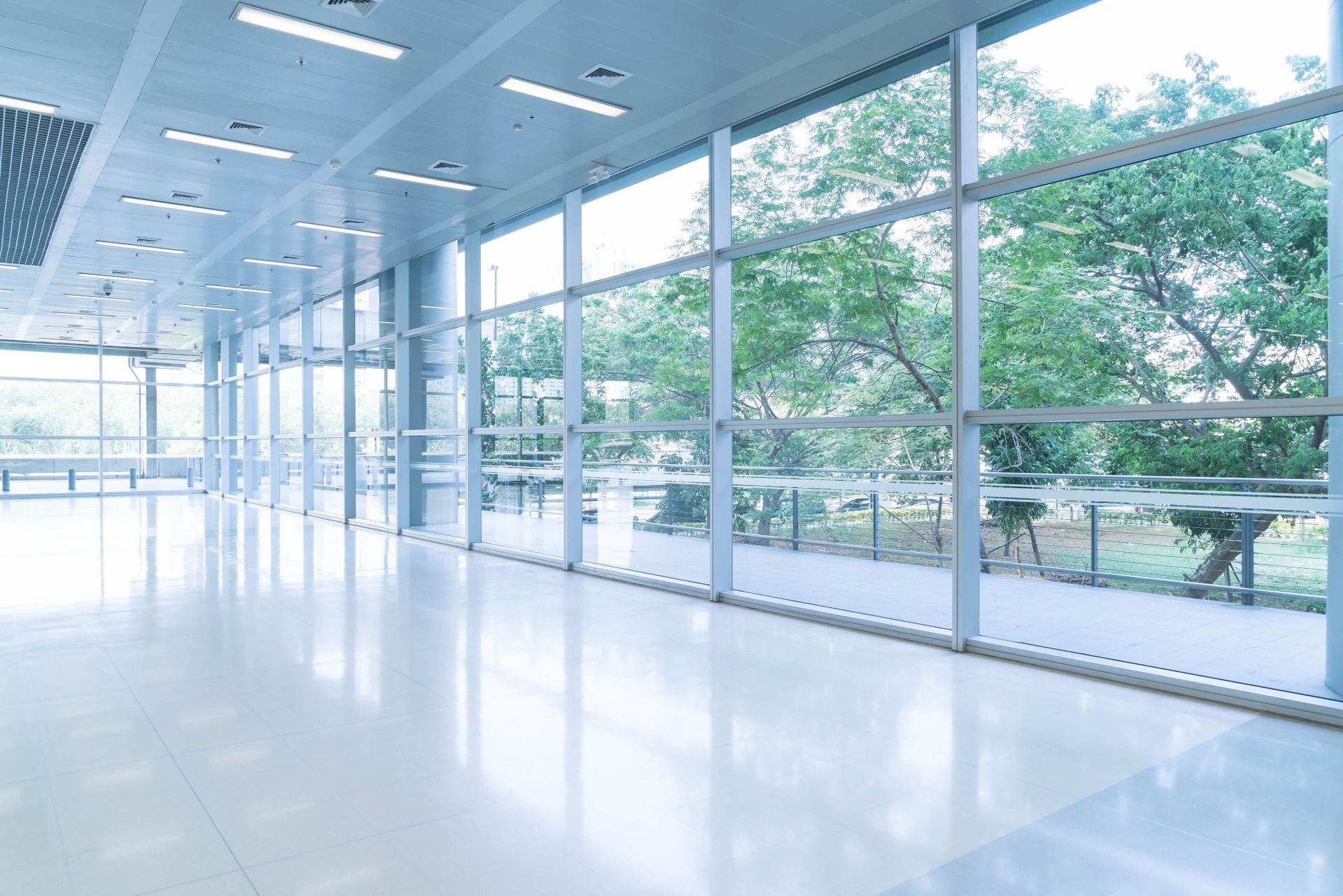 Large, empty modern hallway with floor-to-ceiling windows overlooking trees and outdoor seating.