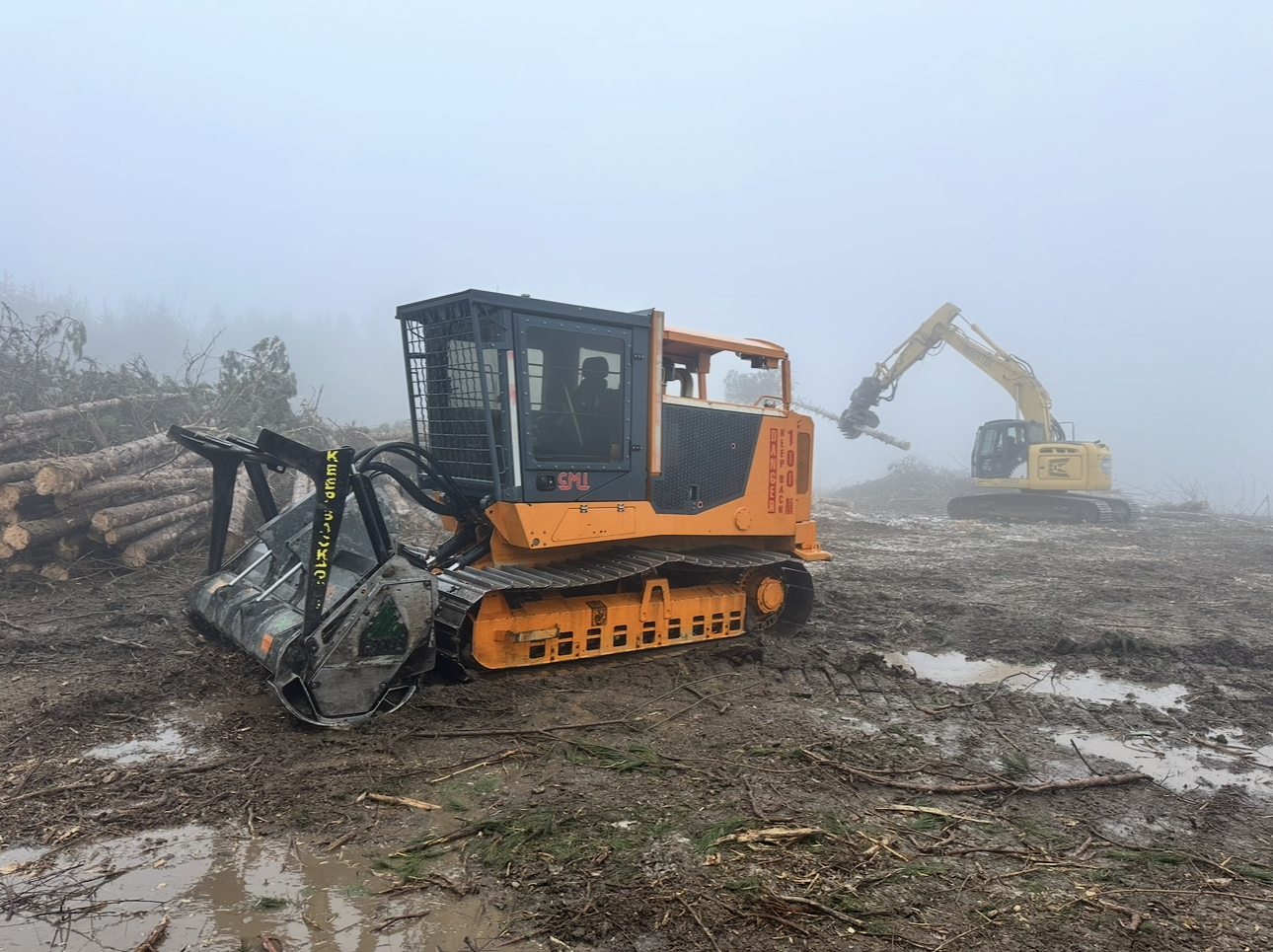 Orange forestry machine on tracks in muddy terrain, with an excavator in the background amidst fog.