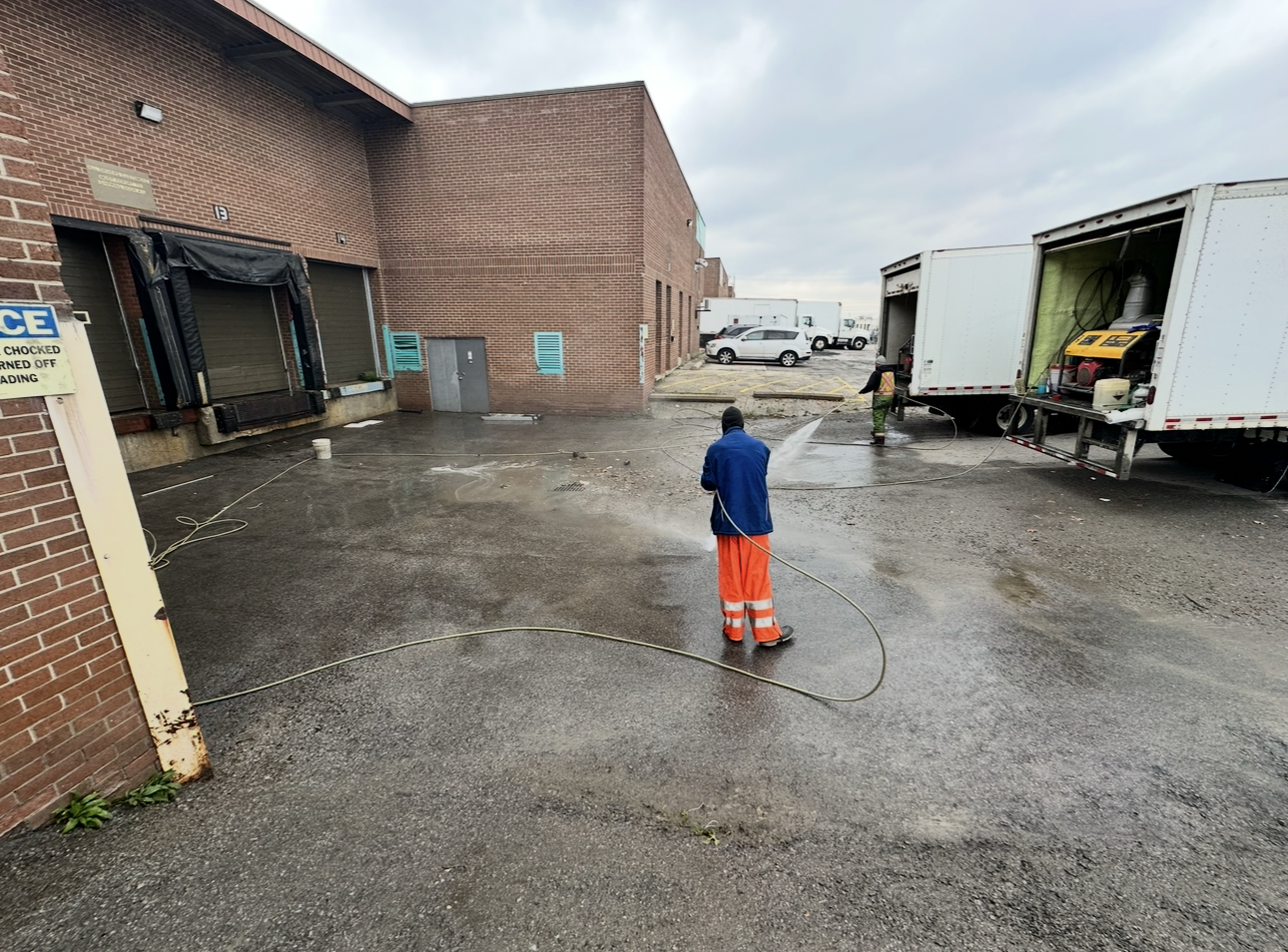 A worker in orange pants sprays water on the wet pavement in front of a brick building and two trucks.