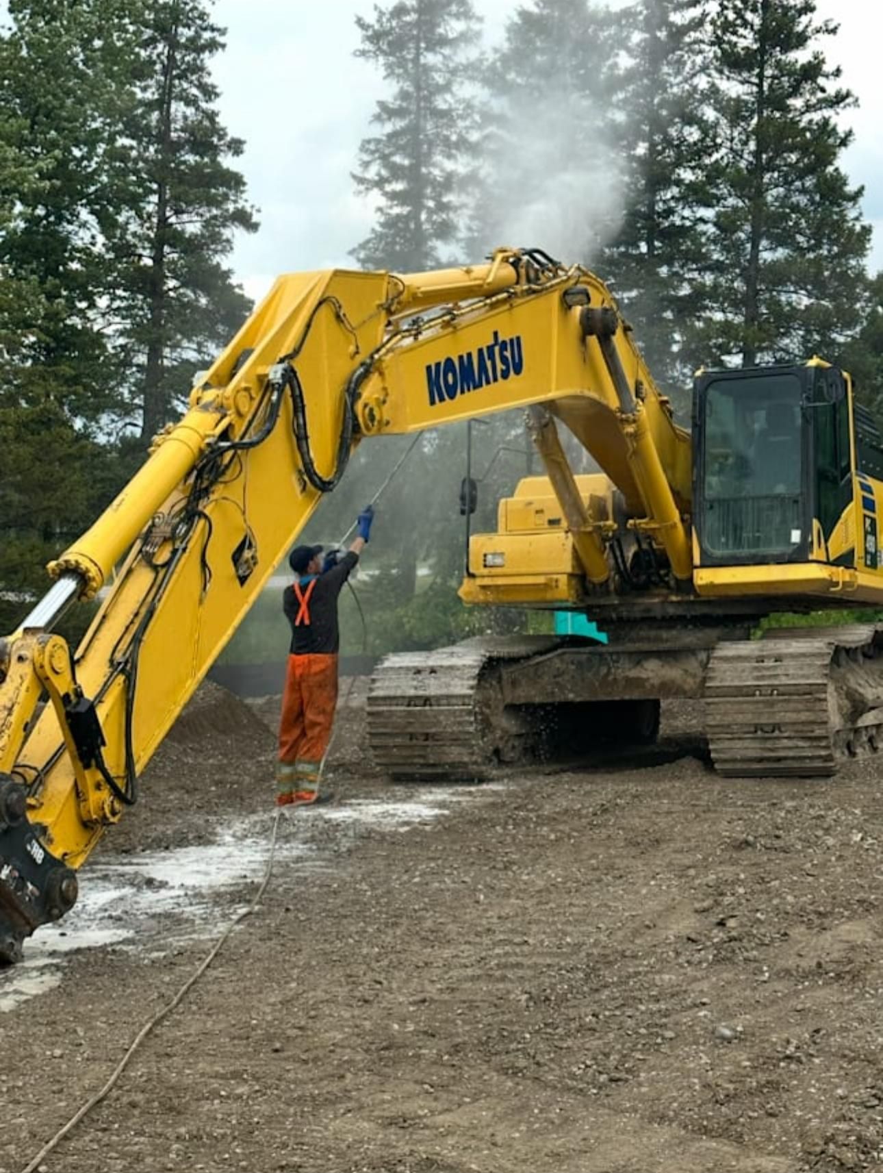 A worker in orange coveralls washes a large yellow Komatsu excavator with a pressure washer; trees in background.