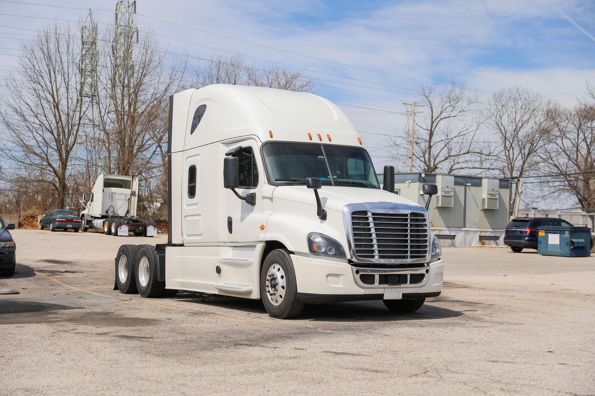 White semi-truck parked in a lot on a sunny day. Another truck and cars are in the background.