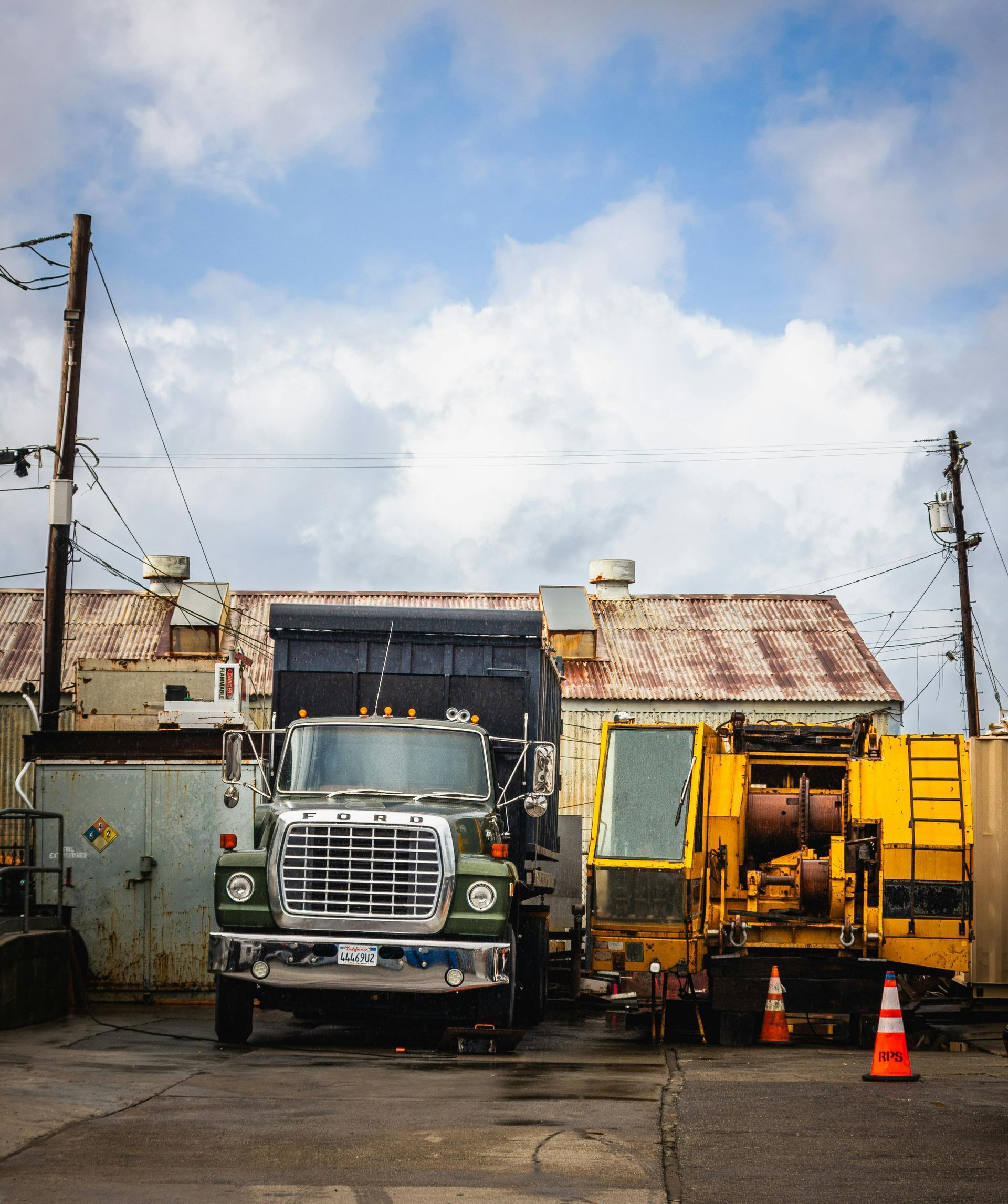 Green dump truck and yellow construction vehicle parked in front of a weathered building under a cloudy sky.