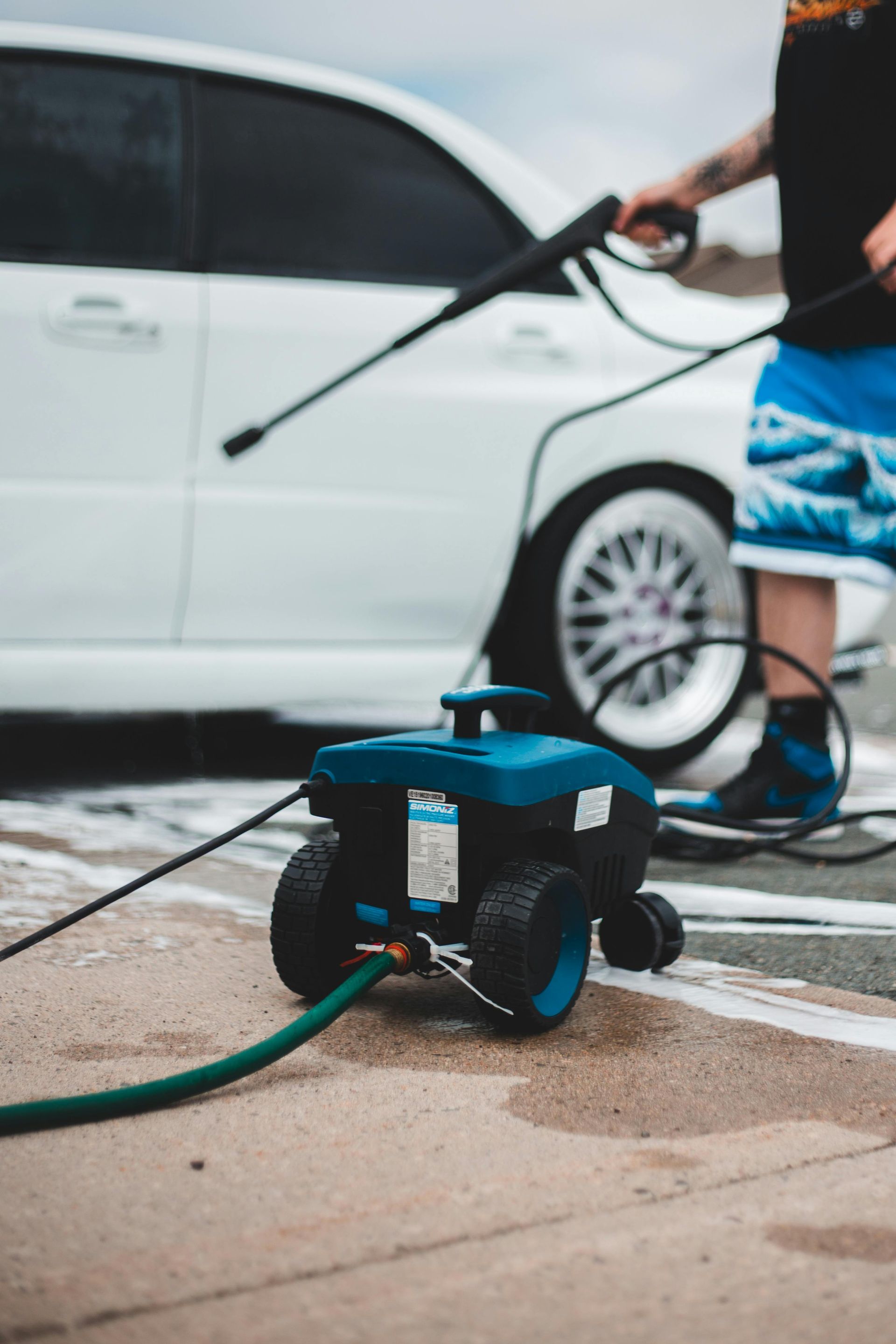 Person washing a white car with a blue pressure washer outside, soapy.
