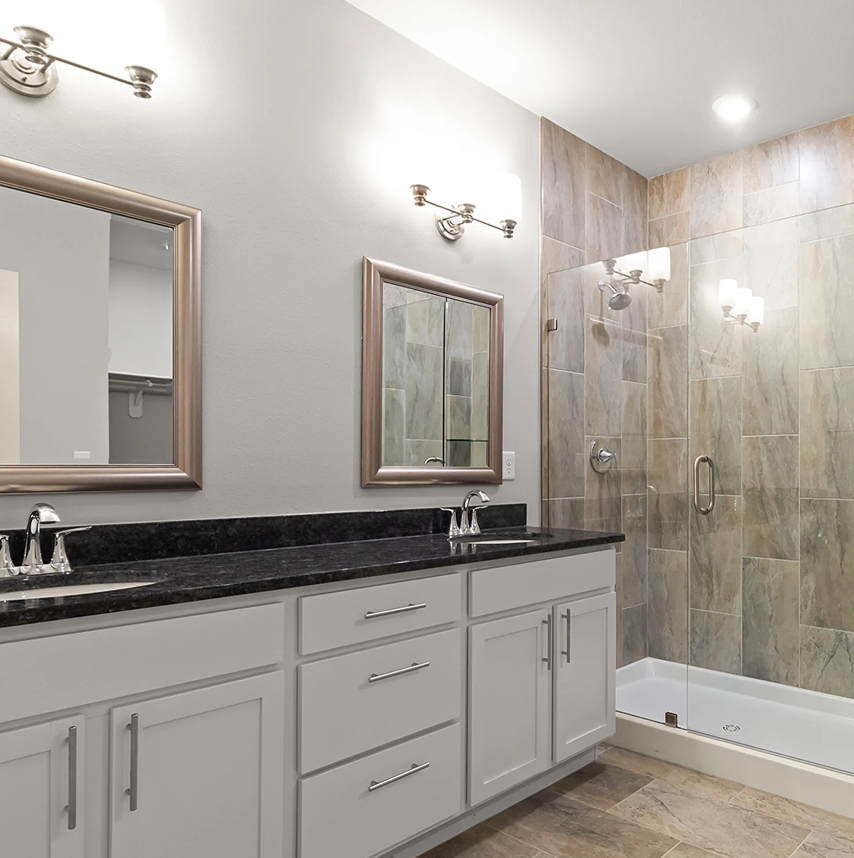 Bathroom with dual sinks, mirrors, shower, and gray walls, tiled flooring and accent wall.