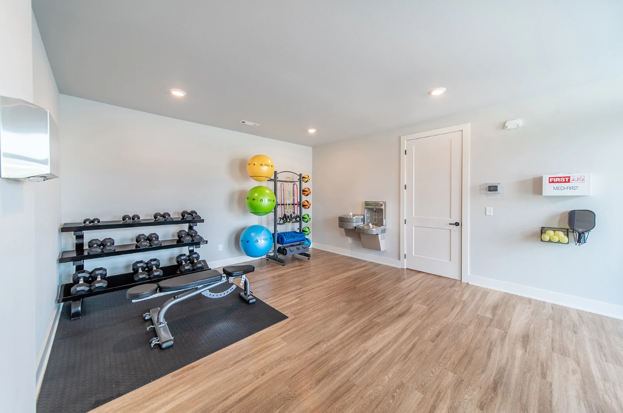 Gym with weights, bench, exercise balls, water fountain, and first-aid kit. Light wood-look flooring and light gray walls.