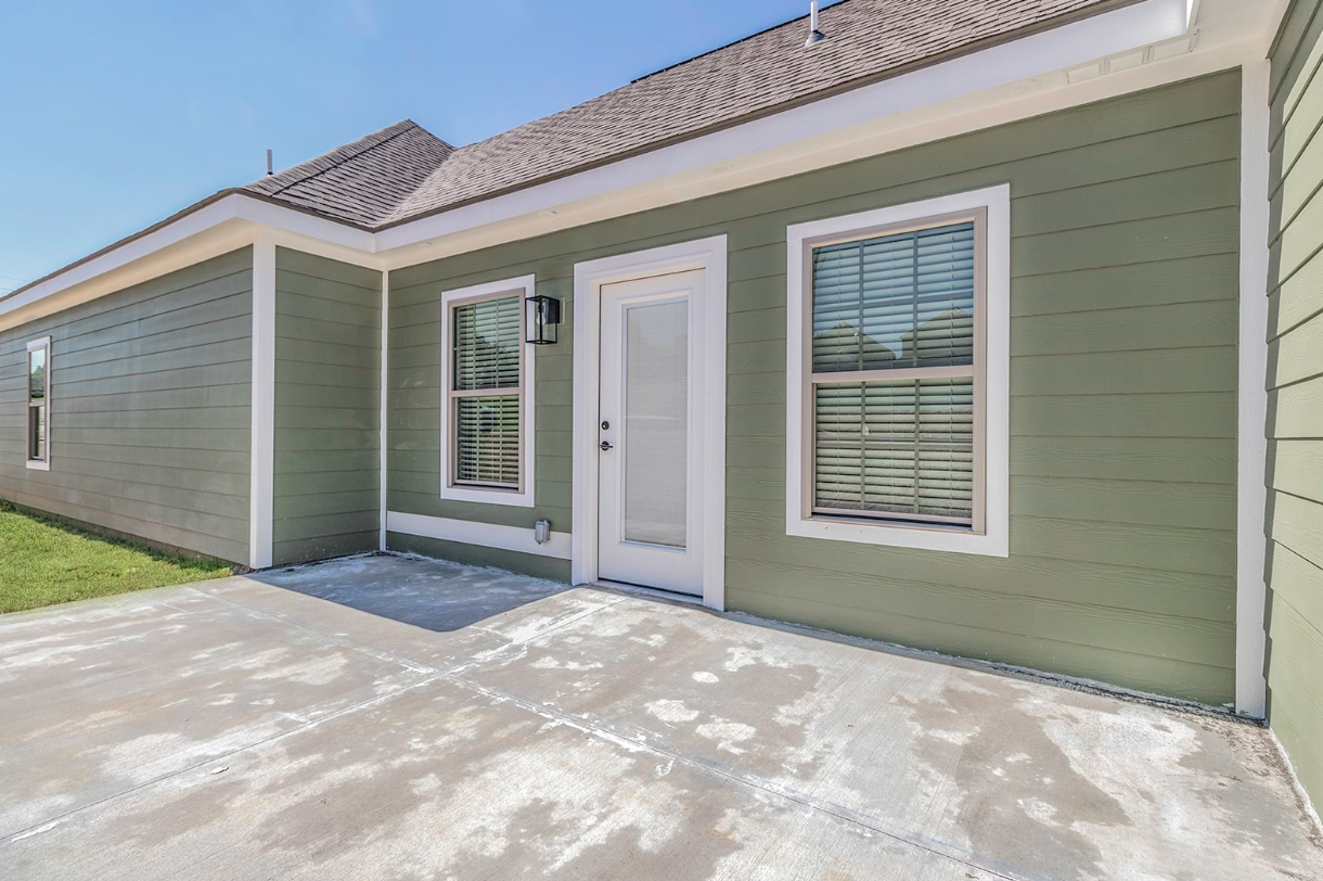 Back patio of a house with green siding, a glass door, windows, and concrete flooring.
