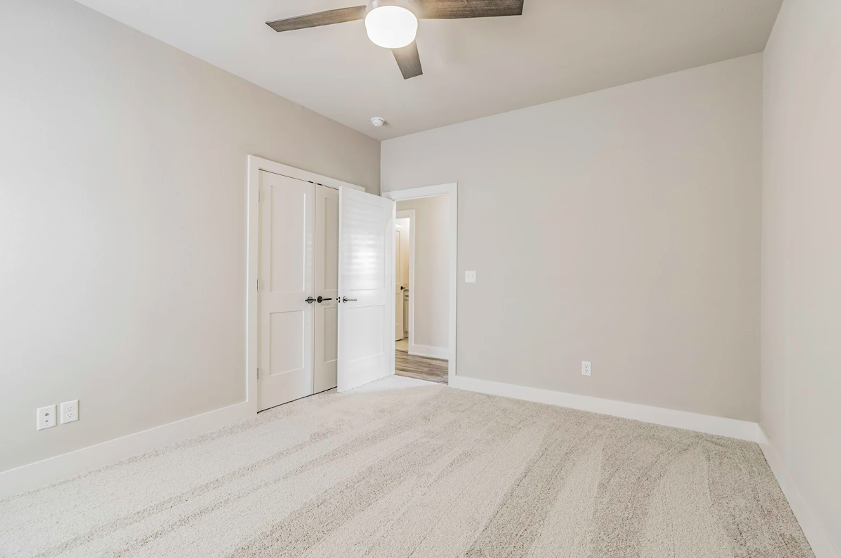 Empty bedroom with beige carpet, white walls, and a ceiling fan.