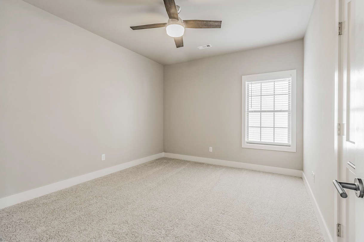 Empty bedroom with tan carpet, a window, ceiling fan, and beige walls.
