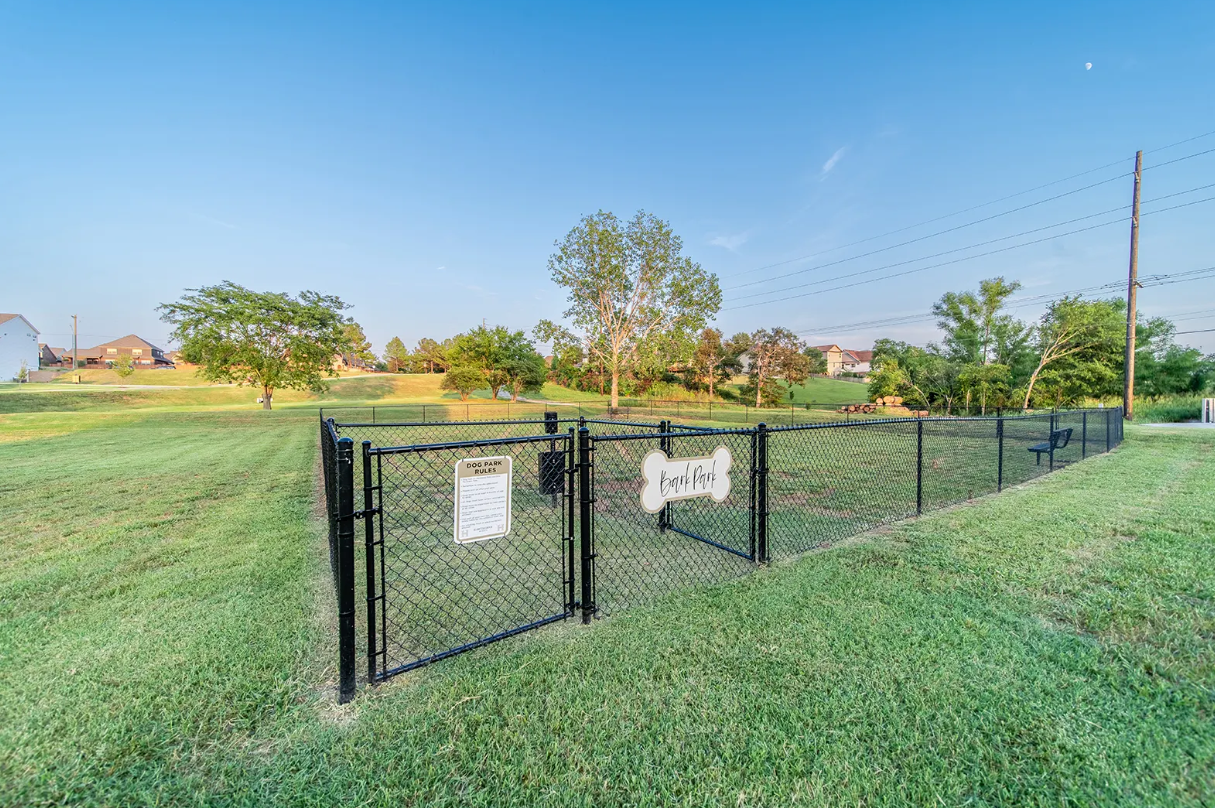 Dog park with black fence, open gate, and sign on a grassy field under a blue sky.