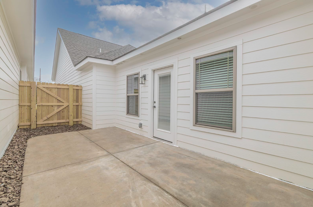 Concrete patio with a white house, door, windows, and wooden fence. Cloudy sky.